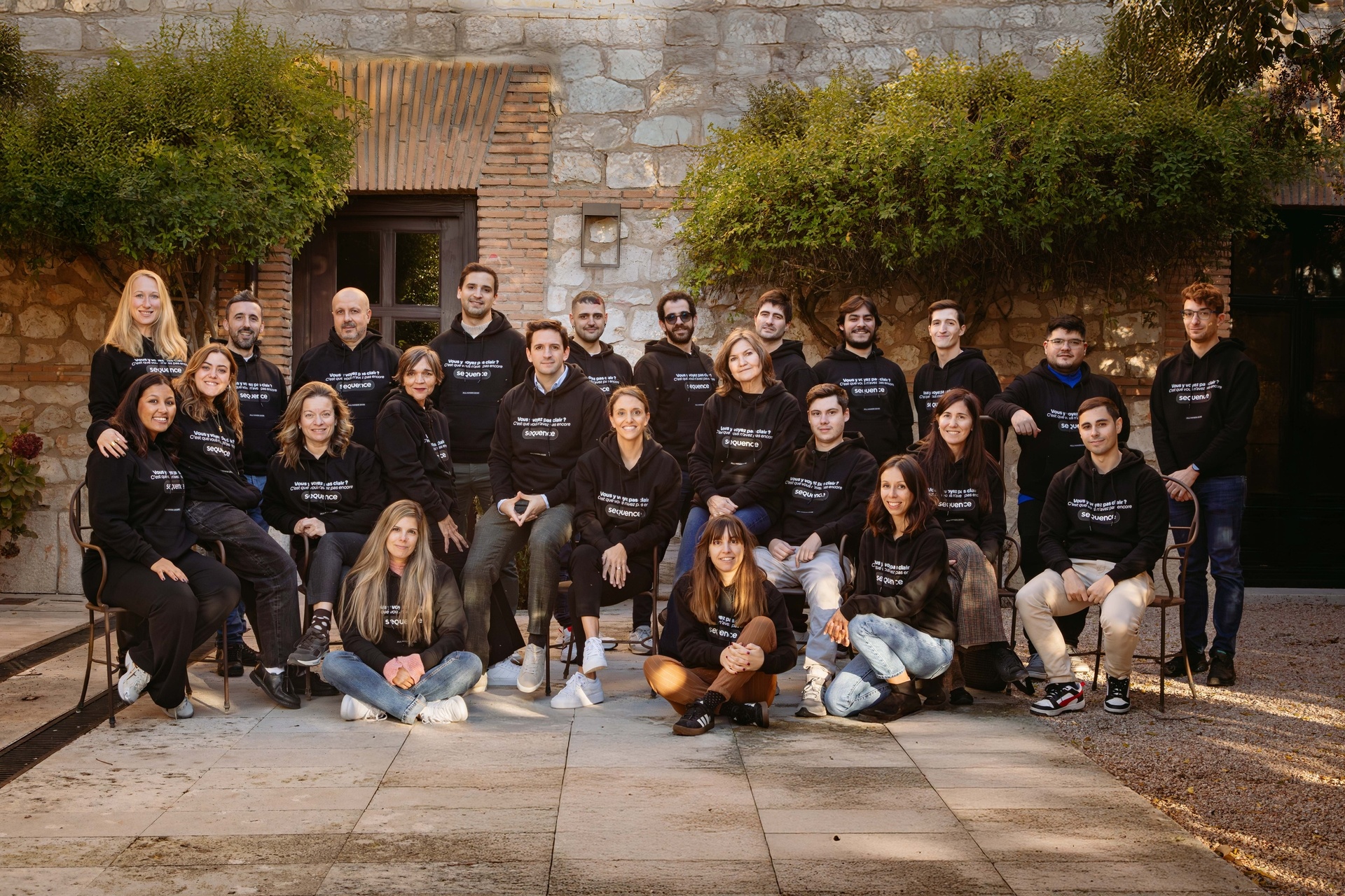 A group photo of a diverse team posing together outside a stone building, all wearing matching black hoodies.