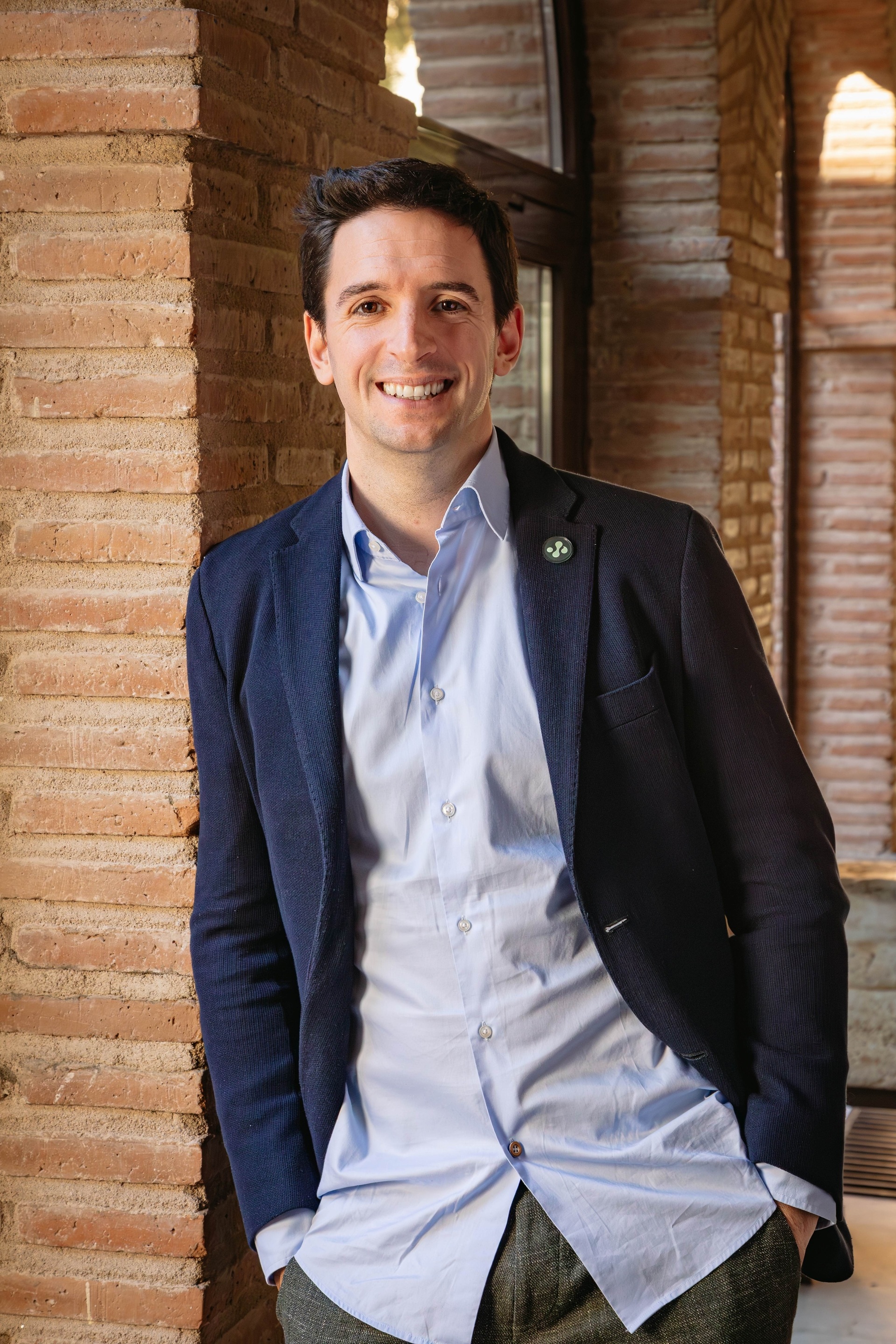 A smiling man in a casual outfit poses against a brick wall, showcasing a relaxed demeanor.